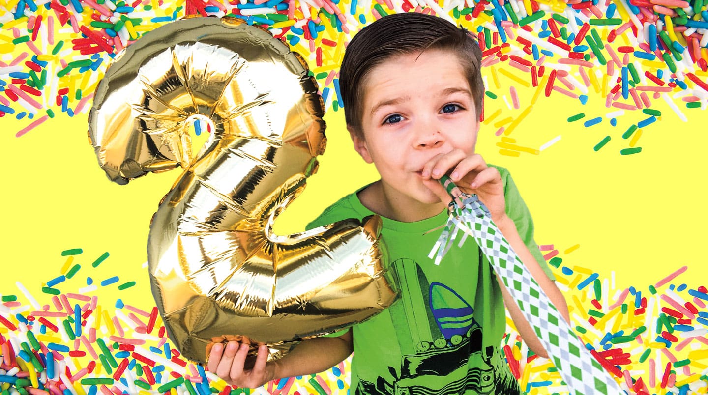 A young boy blows into a noise maker and holds a balloon shaped as the number 2.