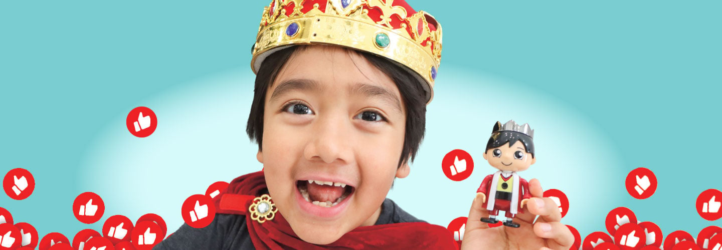 A young boy smiles while wearing a crown and holding a small toy that looks like him.