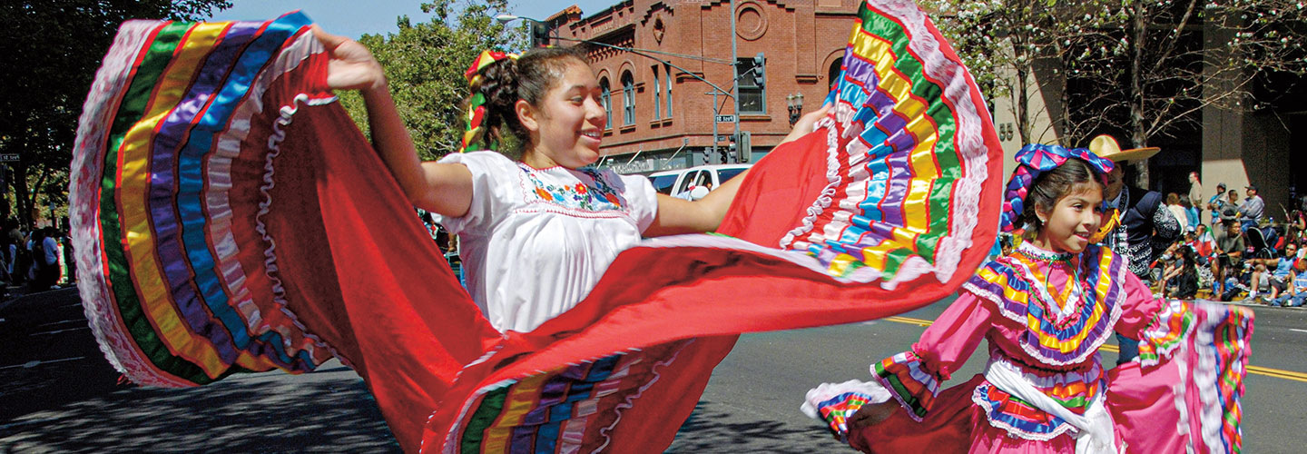 Girls wear traditional Mexican dresses while dancing in a parade.