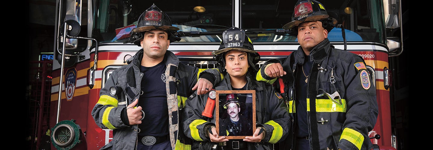 Three firefighters in gear stand before a fire truck as one holds a framed portrait of a firefighter
