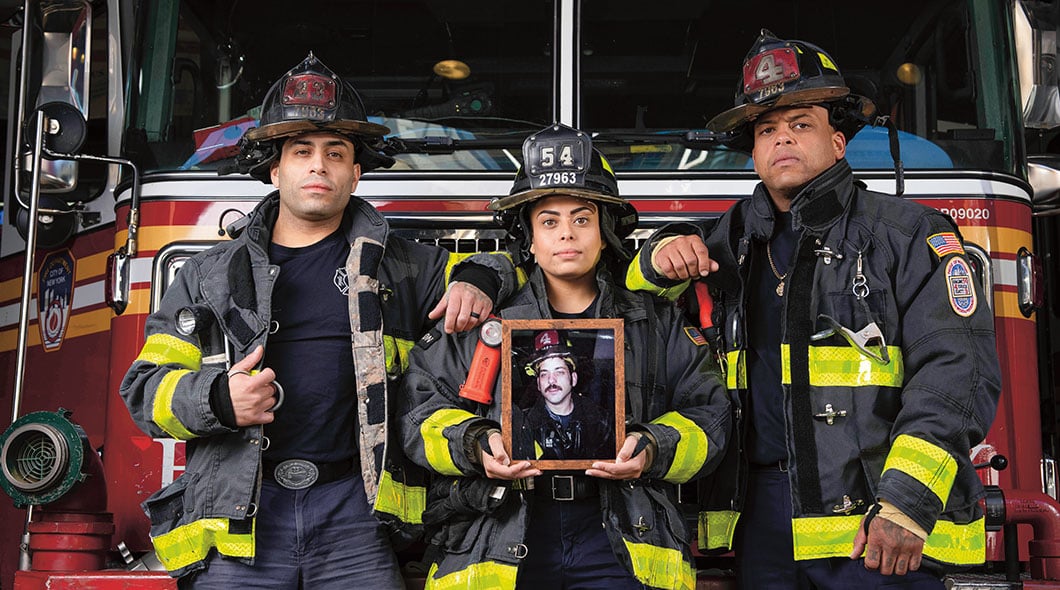 Three firefighters stand before a fire truck as one holds a framed portrait of a fellow firefighter