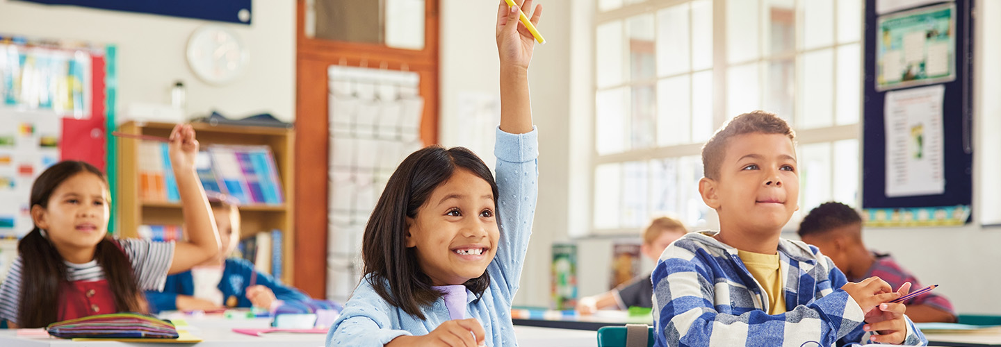 A girl raises her hand at a classroom desk while other students sit nearby