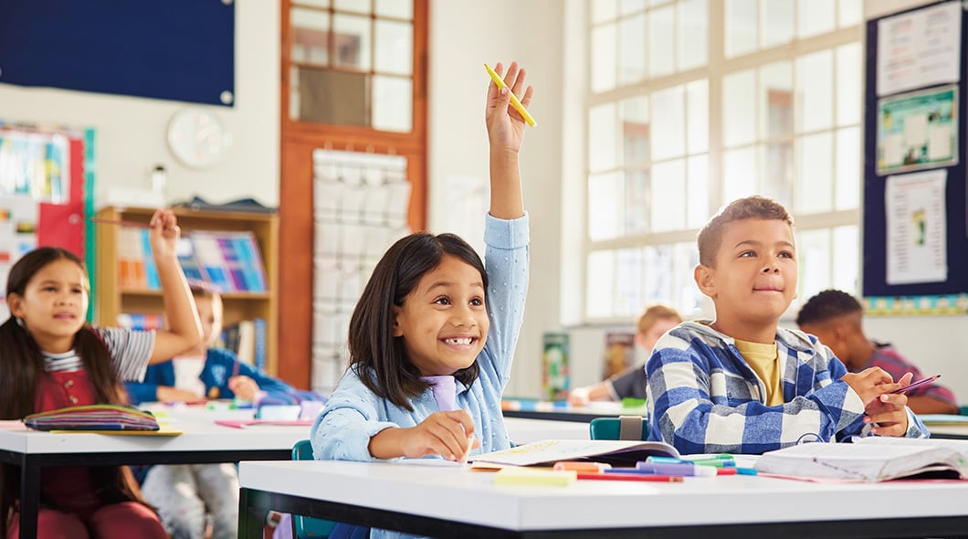 A group of students sit at desks in a classroom and raise their hands to answer a question