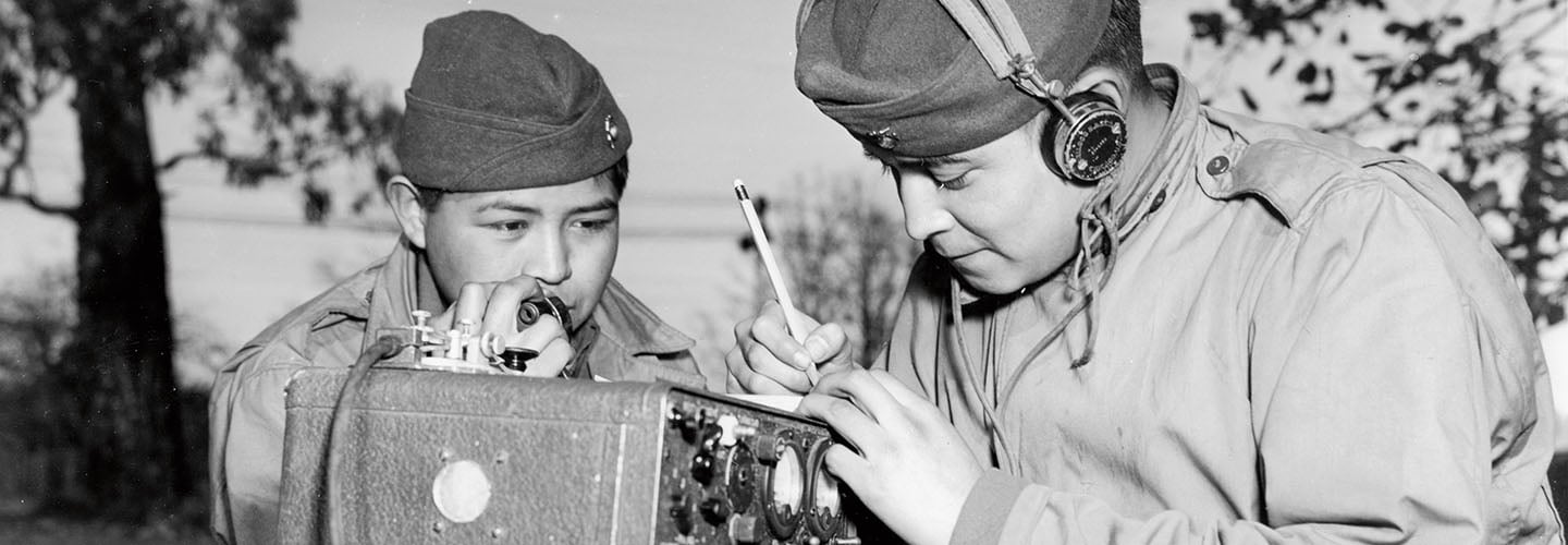 Historical photo of two soldiers in uniform, one speaks into a field radio as the other writes notes