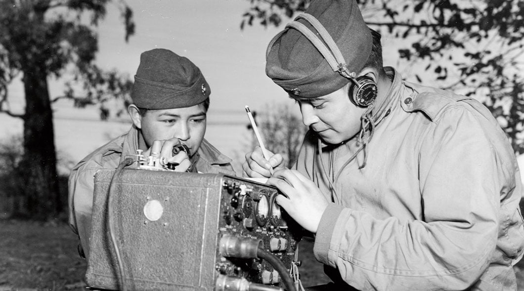 Historical photo of a soldier speaking into a field radio handset while another writes with a pencil
