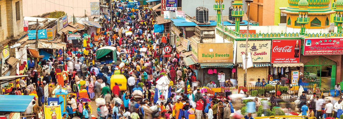 A dense crowd fills a busy market street lined with shops and colorful signs in an Indian city