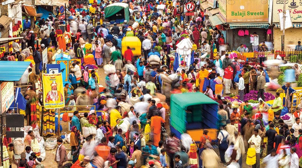 A dense crowd fills a busy outdoor market street with vendors, flowers, and auto-rickshaws